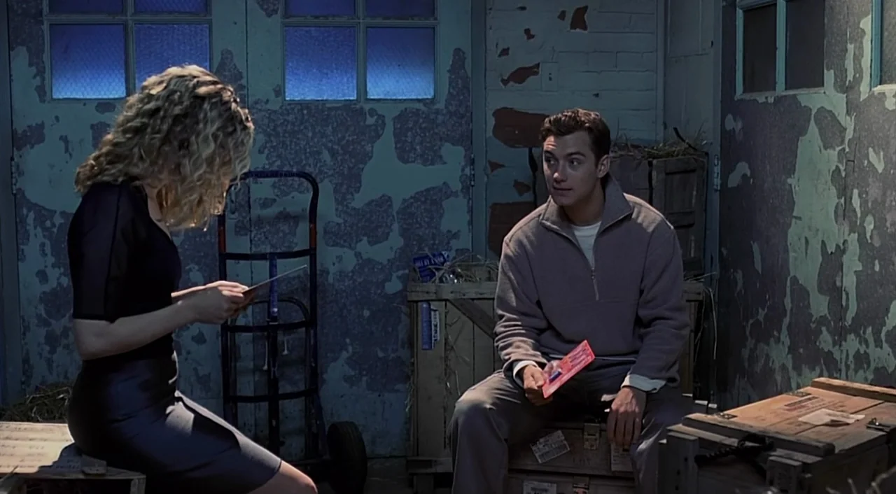 eXistenZ (1999) movie still: medium — A cluttered storage space features distressed blue paint peeling from brick walls. A woman and a man sit on…