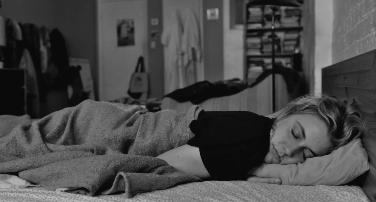 Frances Ha (2012) movie still: medium — A furnished room includes a wooden bed headboard, a bookshelf, and a door. A young woman lies horizontally…