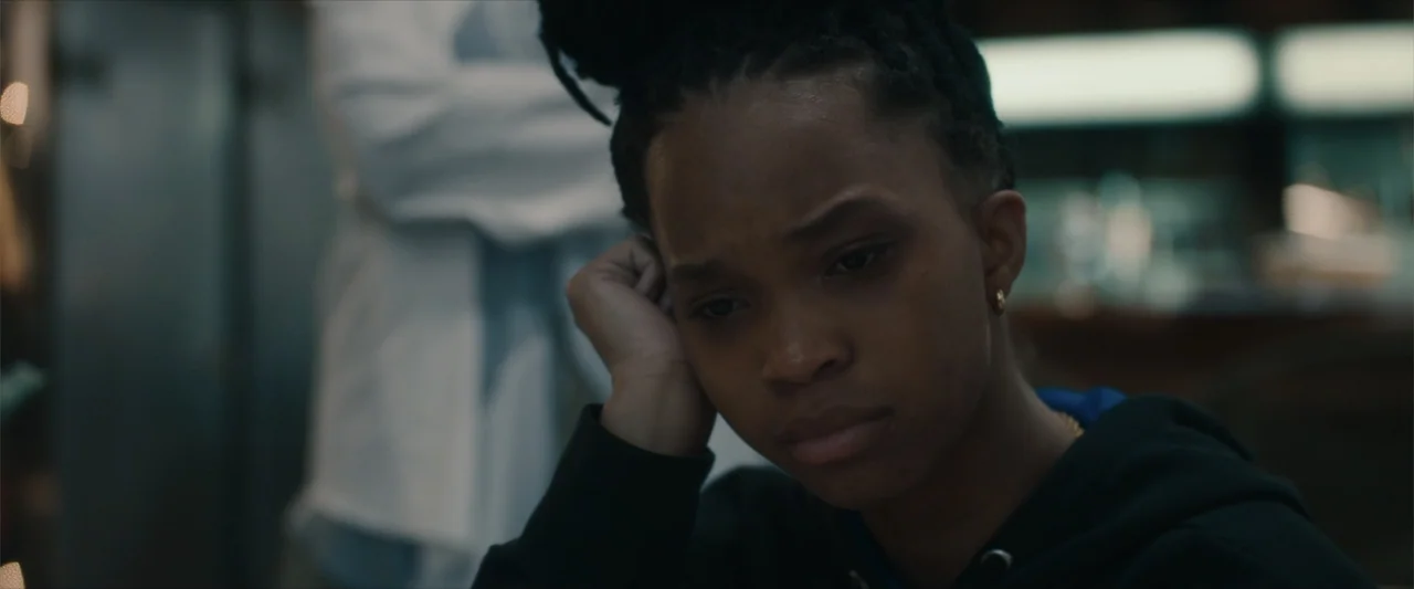 Breathe (2011) movie still: closeup — A dark indoor space with a blurred person in a white top in the background. A young woman with her hair in an…