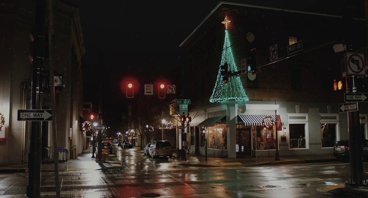 The Wolf of Snow Hollow (2020) movie still: wide — A wet street intersection at night decorated for Christmas with a large light display. A…