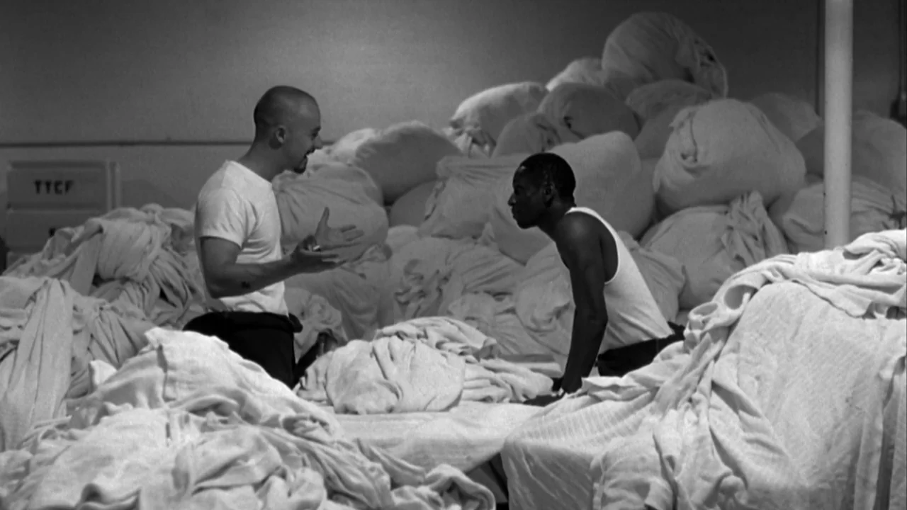 American History X movie still: medium — Two men stand amidst large piles of stacked laundry bags in a facility. One man gesturing toward the other…