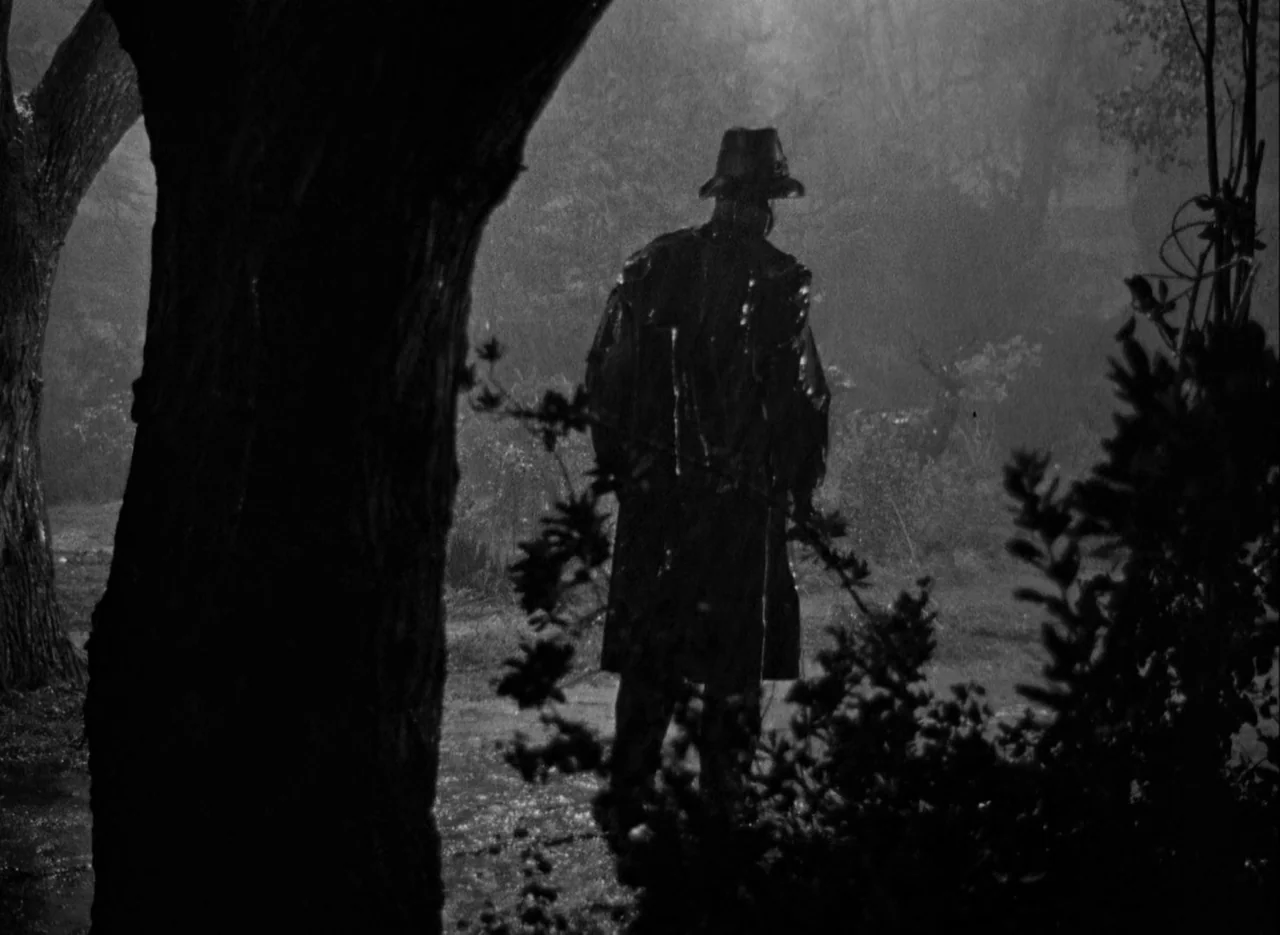The Spiral Staircase (1946) movie still: fullbody — A rain-drenched park at night features dark trees and wet ground. A man in a trench coat and…