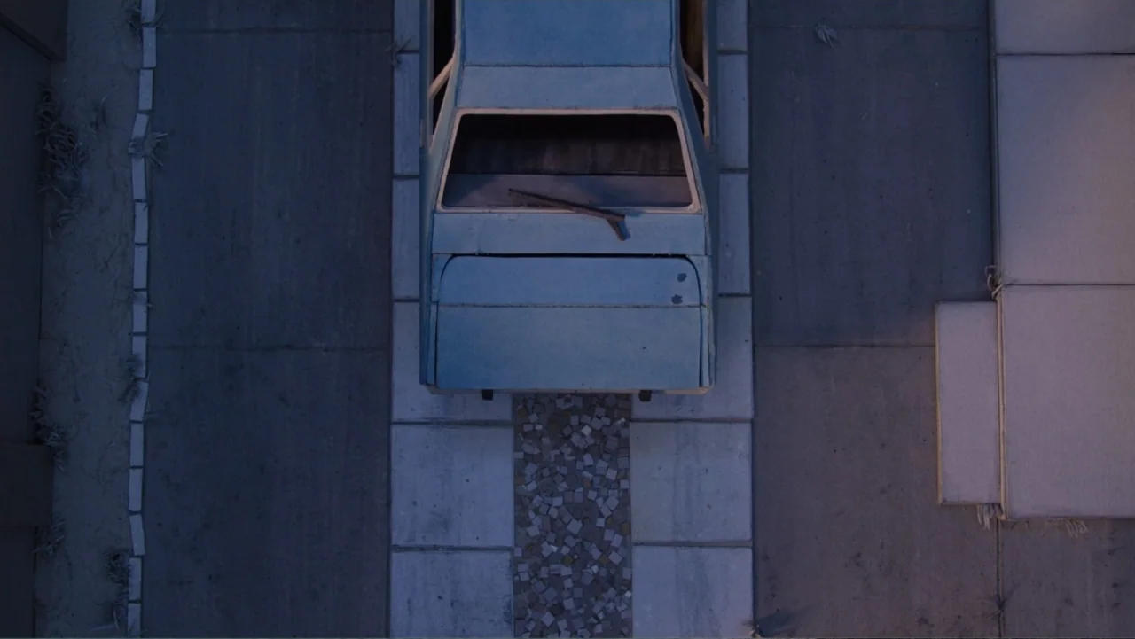 Bestia (2017) movie still: establishing — A top-down view of a light blue sedan parked on a concrete residential driveway. The car is centered on a…