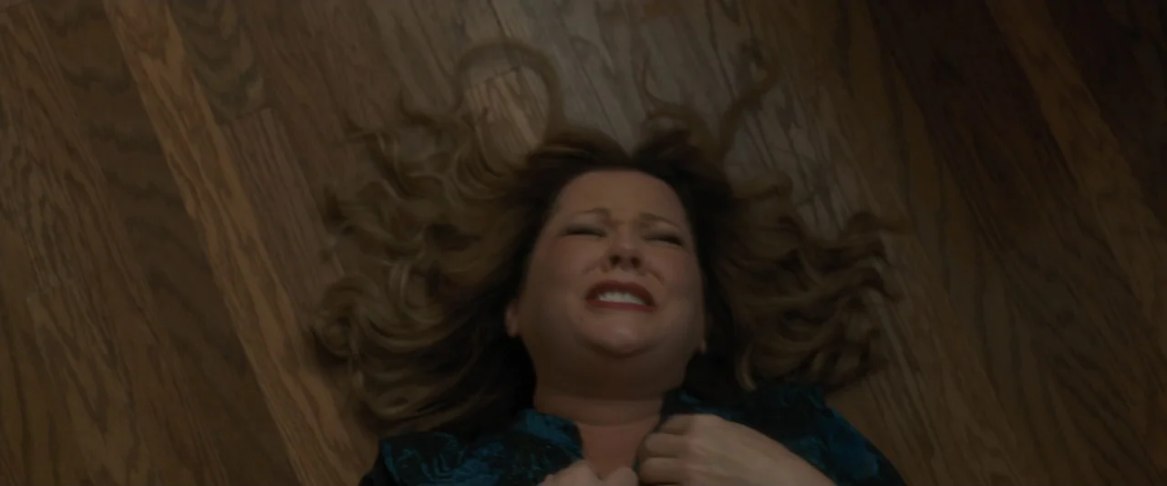 Life of the Party (2018) movie still: closeup — A dark, plain interior setting featuring a woman lying on a dark hardwood floor. The subject is on…