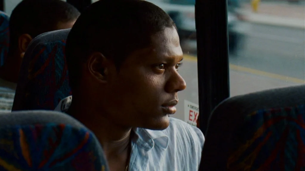 Sugar (2008) movie still: medium — The interior of a public bus with patterned fabric seat headrests. A young man sits in a seat looking out the…