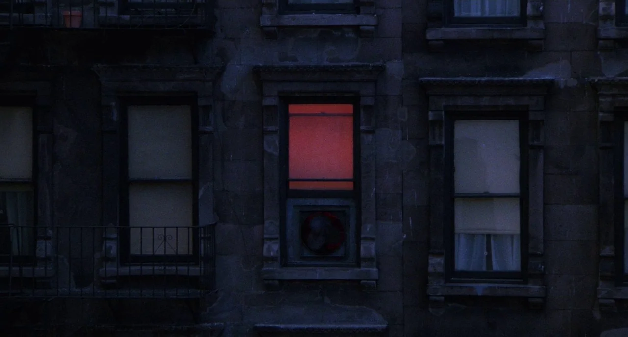 Angel Heart (1987) movie still: wide — A dark brick apartment building facade features multiple windows at night. One window emits a distinct red…
