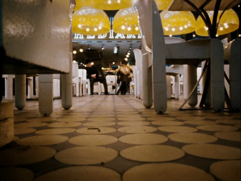 Making of: World On A Wire (1973) movie still: wide — A diner interior features rounded floor tiles and white plastic furniture in the foreground. A…