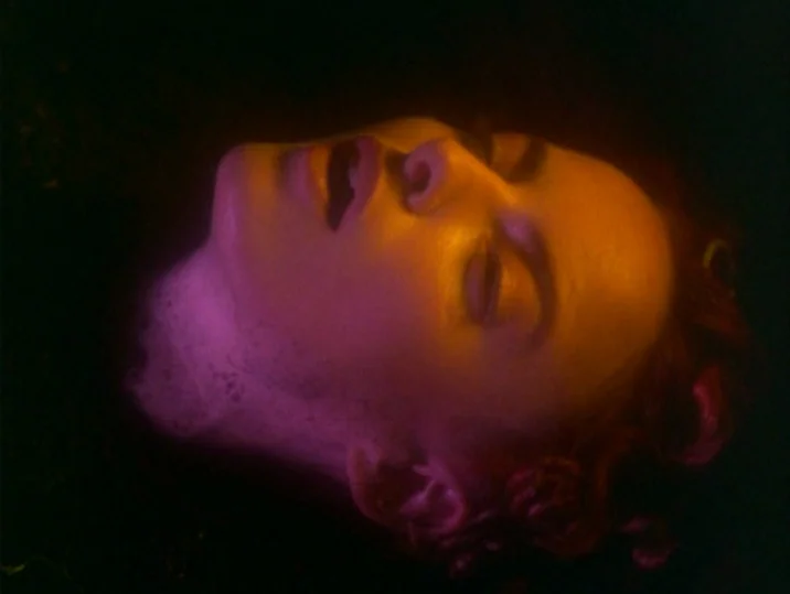 Twilight of the Ice Nymphs (1997) movie still: closeup — A woman's face rests on the dark surface of water at night. Intense colored light reflects…