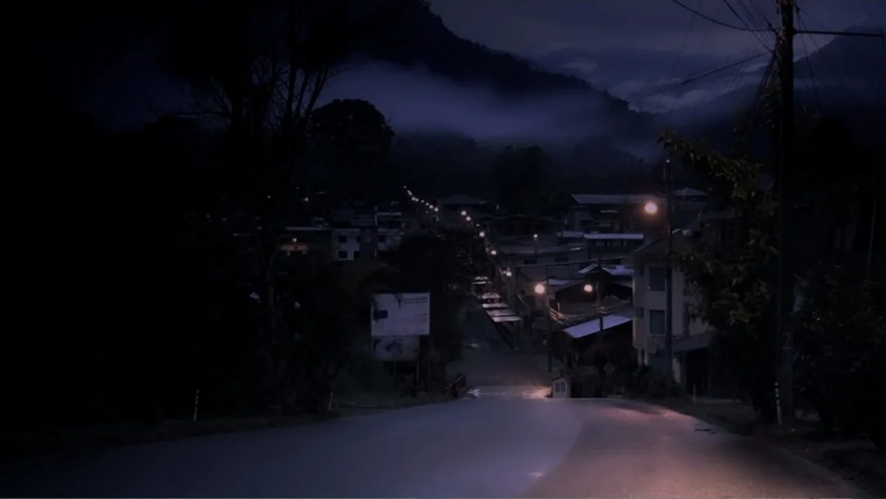 El Duende Sátiro (2016) movie still: establishing — A wet asphalt road slopes downhill into a small, sparsely lit mountain town at night. Low clouds…