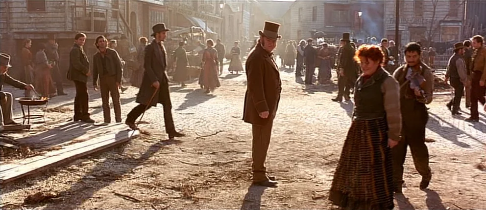 Gangs of New York (2002) movie still: wide — A crowded dirt thoroughfare in a 19th-century frontier town with rustic wooden buildings. Many people…