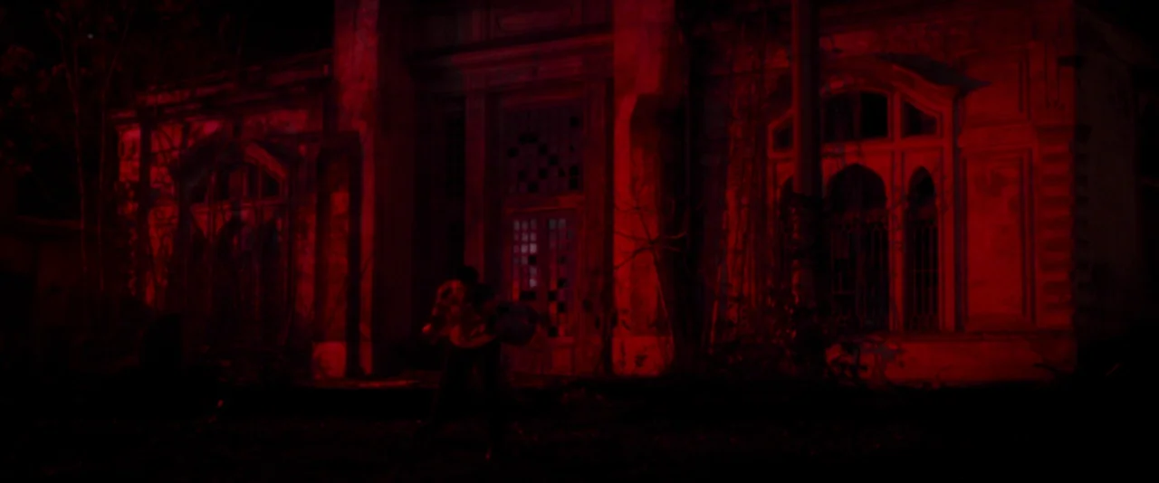 Baskin (2015) movie still: wide — An abandoned manor with overgrown vines stands in deep shadow during the night. A person moves toward the central…