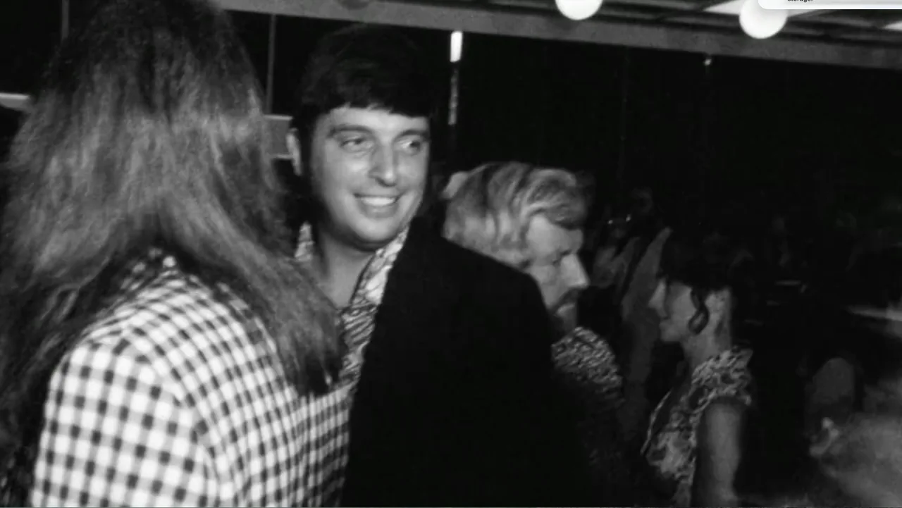 Becoming Led Zeppelin (2025) movie still: medium — A crowded interior event space features multiple people in 1970s formal wear. A man with…
