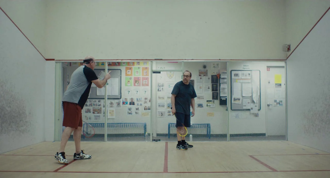 Private Life (2018) movie still: wide — An indoor squash court features wooden floors and high walls covered in posters and notices. Two middle-aged…