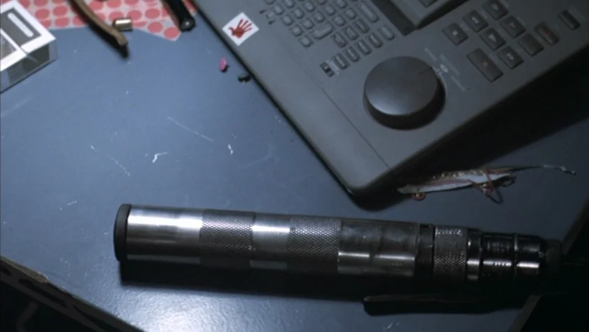 Benny’s Video (1992) movie still: closeup — A dark blue desk surface contains a silencer, an editing controller, and small accessories. The editing…