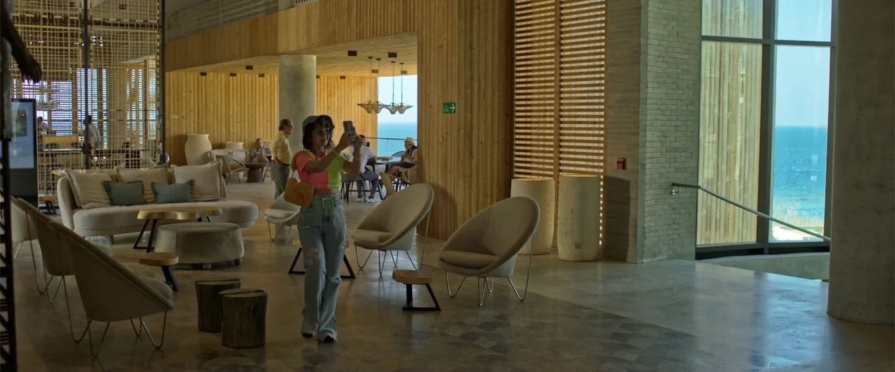 Férias Trocadas (2024) movie still: fullbody — A spacious resort lobby features light wood paneling, modern beige furniture, and coastal views. A…