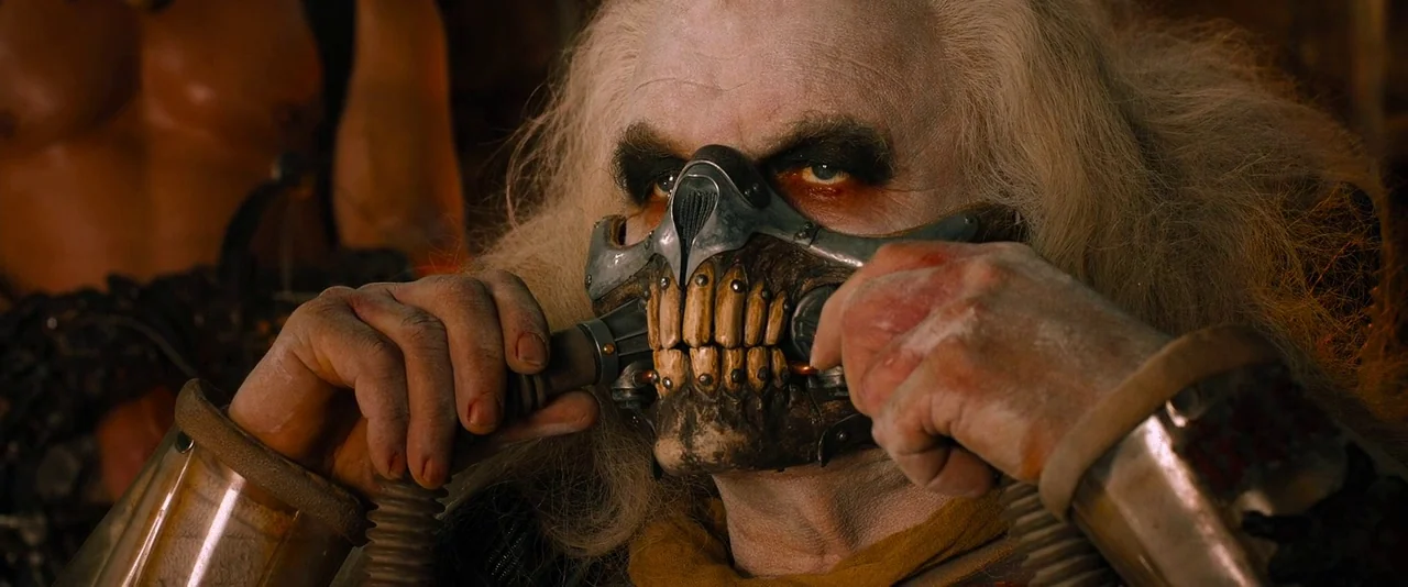 Mad Max: Fury Road (2015) movie still: closeup — A dimly lit industrial environment with metallic machinery and dark shadows visible behind the…