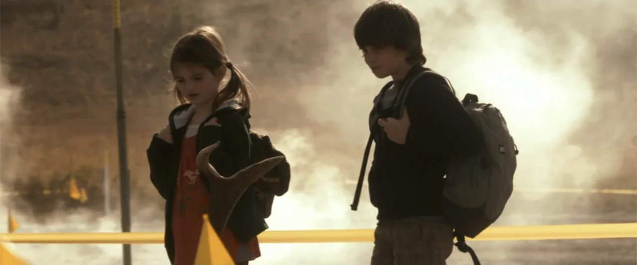 2012 (2009) movie still: medium — A young girl and boy stand in a steam-covered field marked by yellow caution tape. The children carry backpacks…