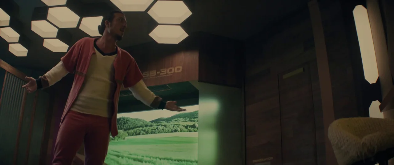 Domain (2017) movie still: medium — A sterile, wood-paneled room features hexagonal ceiling lights and a massive digital scenery screen. A young man…