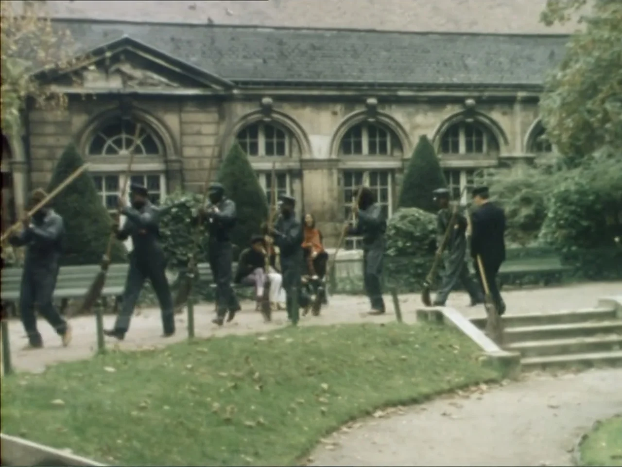 A Dessert for Constance (1981) movie still: wide — A group of workers carries long-handled brooms across a gravel path in a public park. The men…