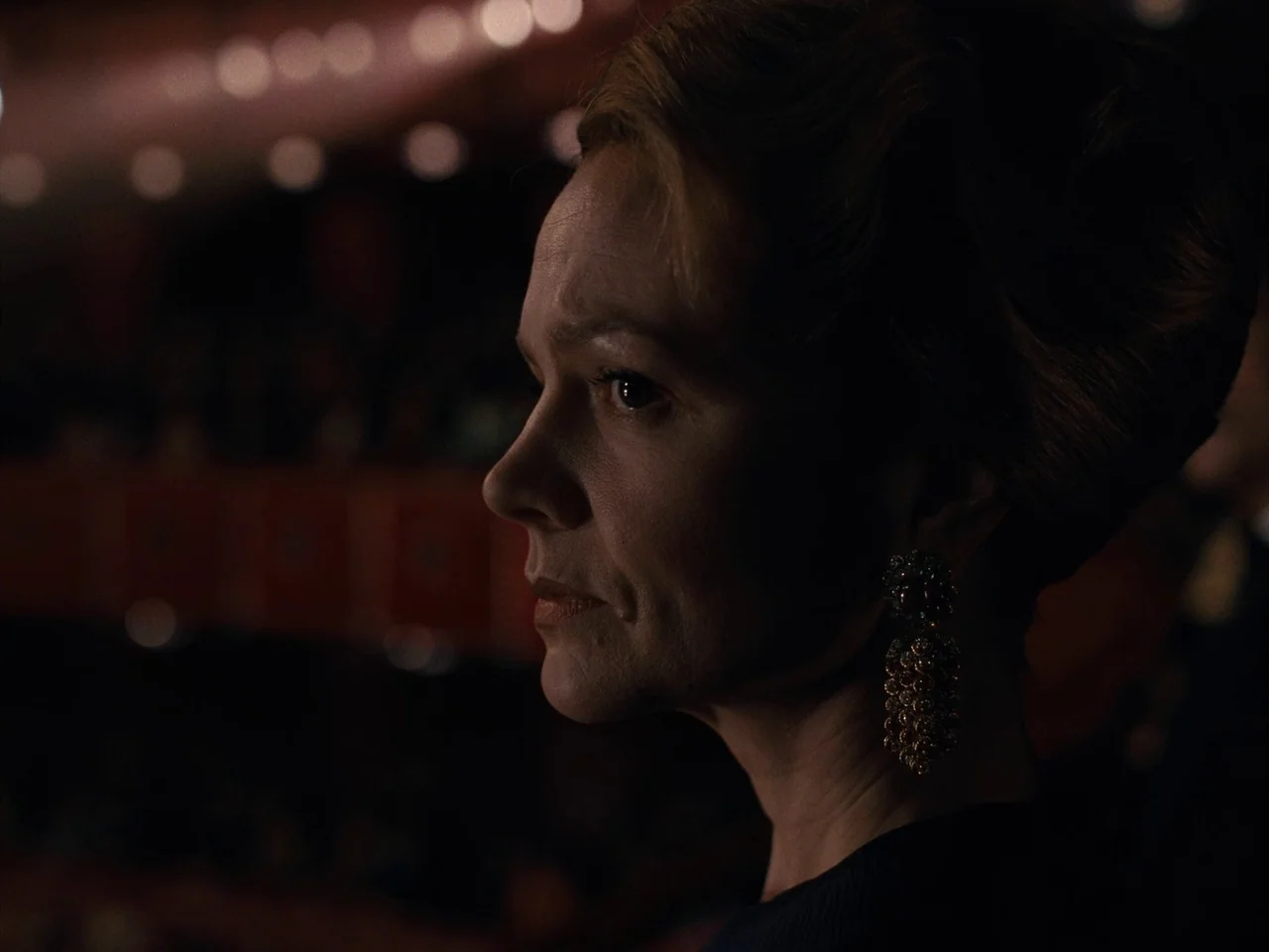 Maestro (2023) movie still: closeup — An interior view of a dimly lit, plush seating area during a public performance. A woman with an elaborate…