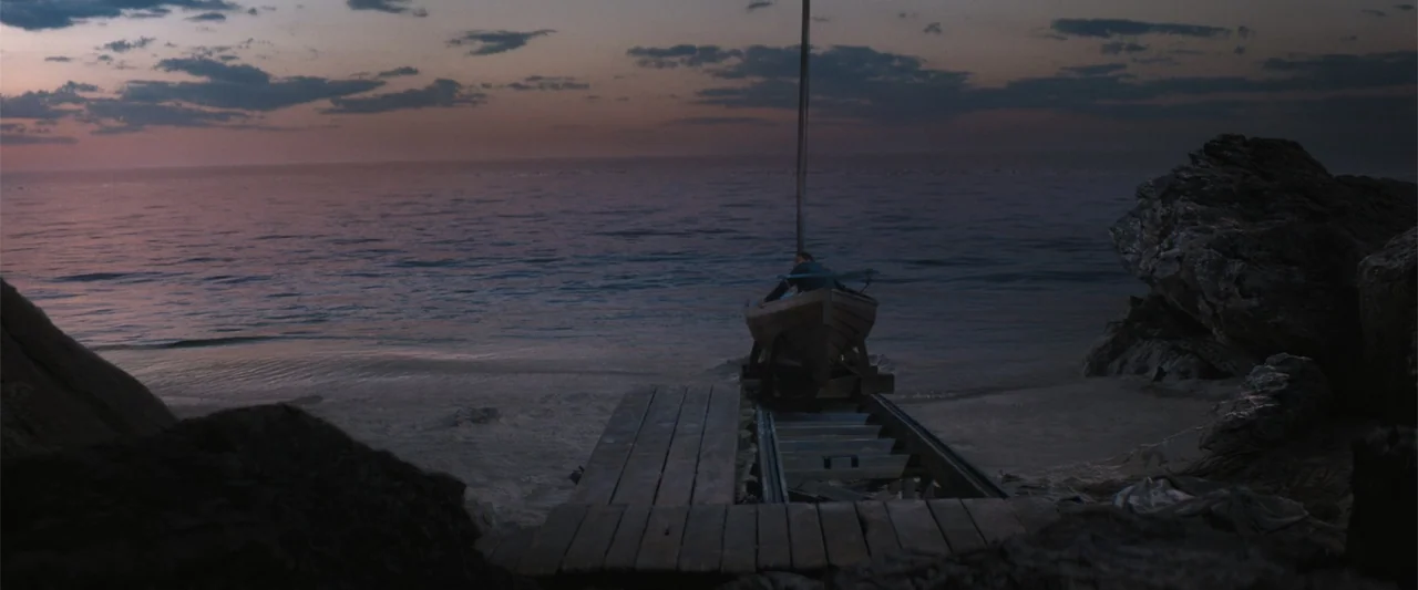 The Bluff (2026) movie still: wide — A wooden boat sits on a rail track leading into a calm, dark ocean. A lone person sits inside the small vessel…