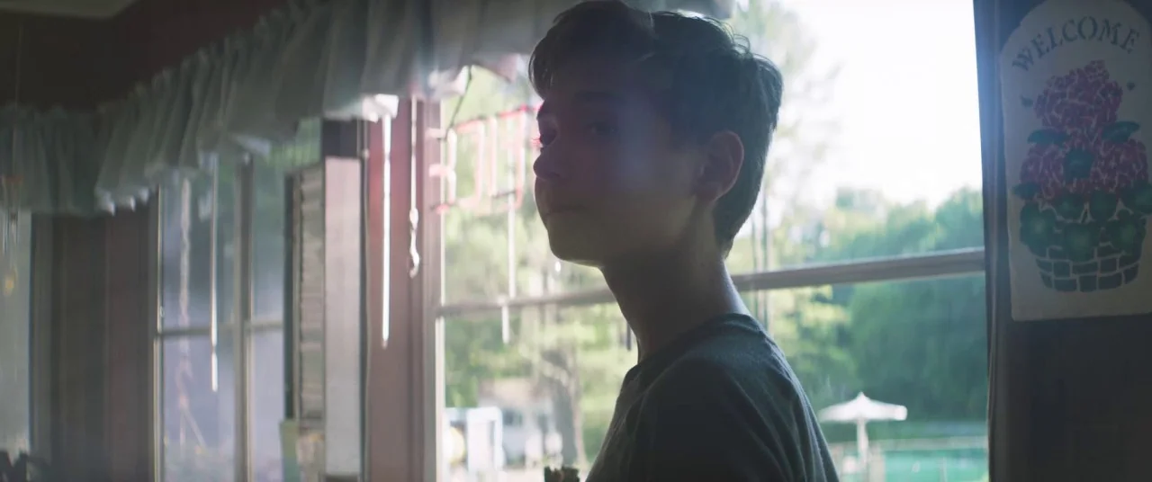 The Strange Ones (2017) movie still: medium — A view inside a sunlit restaurant area with window curtains and a welcome sign. A teenage boy stands…