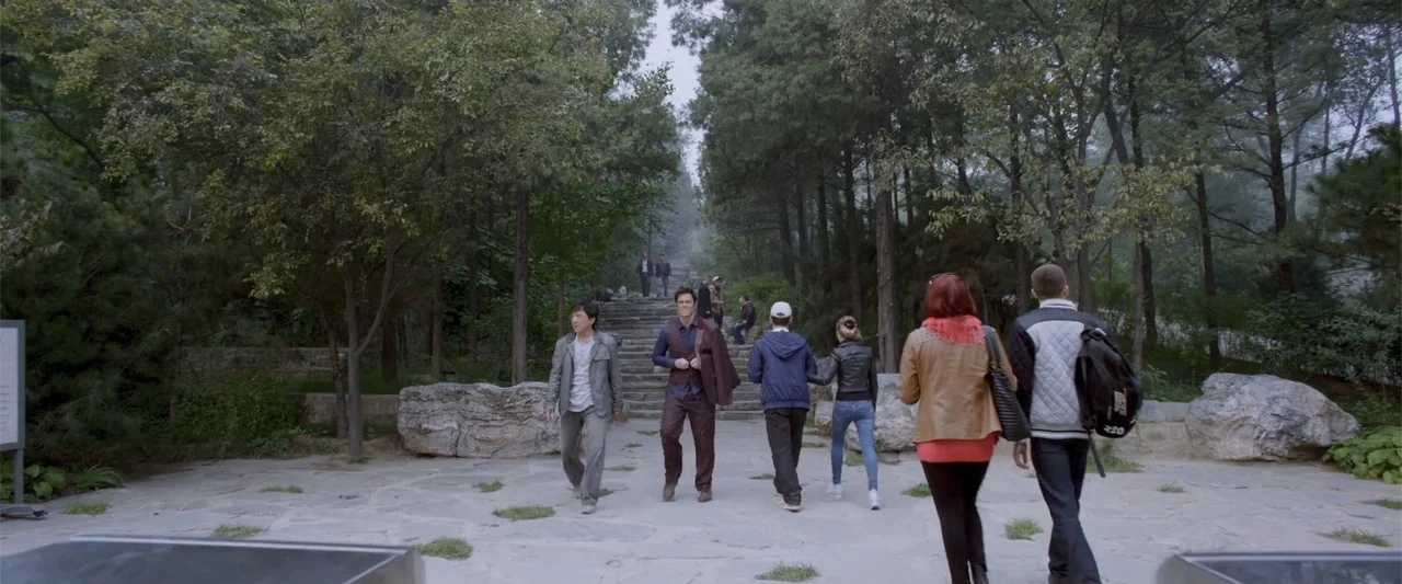 Skiptrace (2016) movie still: fullbody — A stone pathway leads up to a staircase surrounded by dense, tall trees. A diverse group of men and women…