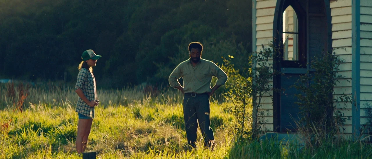 Z for Zachariah (2015) movie still: wide — A man and a woman stand in an overgrown grassy field near a small white shed. The scene is lit by the…