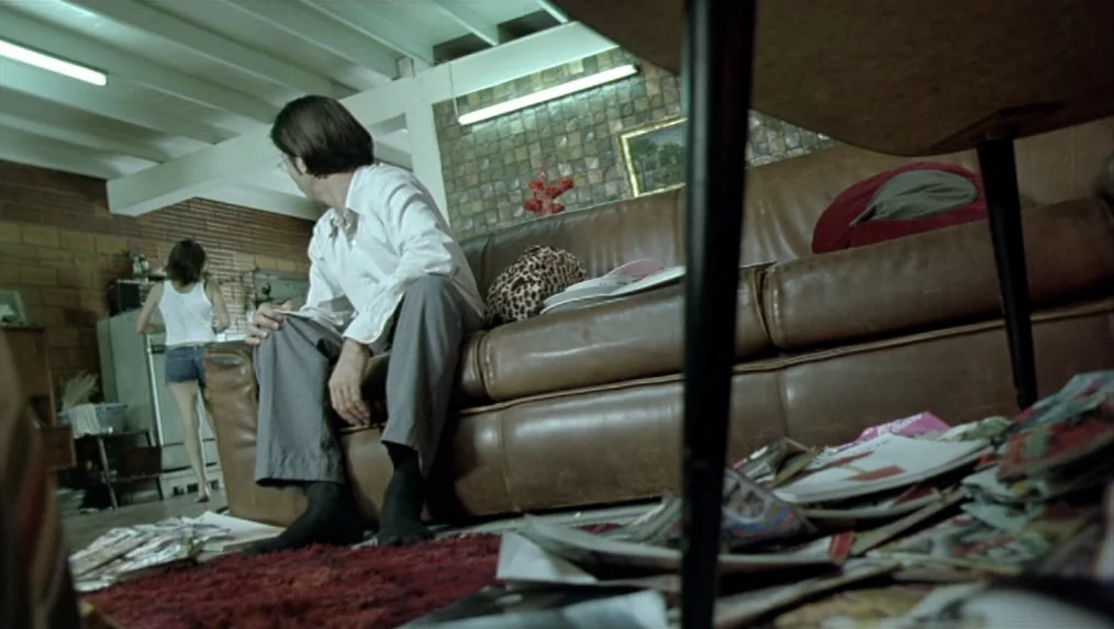 Last Life in the Universe (2003) movie still: medium — A cluttered apartment living room features a leather sofa and a patterned brick wall. A man…
