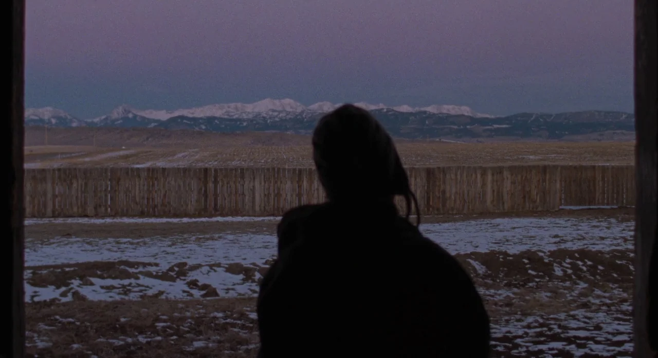 Certain Women (2016) movie still: medium — A rural ranch landscape features a wooden fence line and snow-dusted ground at dusk. A person in shadow…