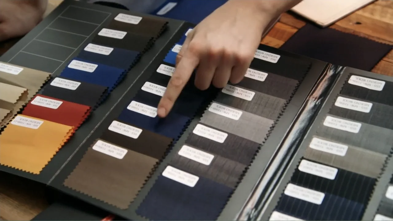 Suited (2016) movie still: closeup — A fabric sample book lies open on a wooden table displaying various textile swatches. A person's hand points…