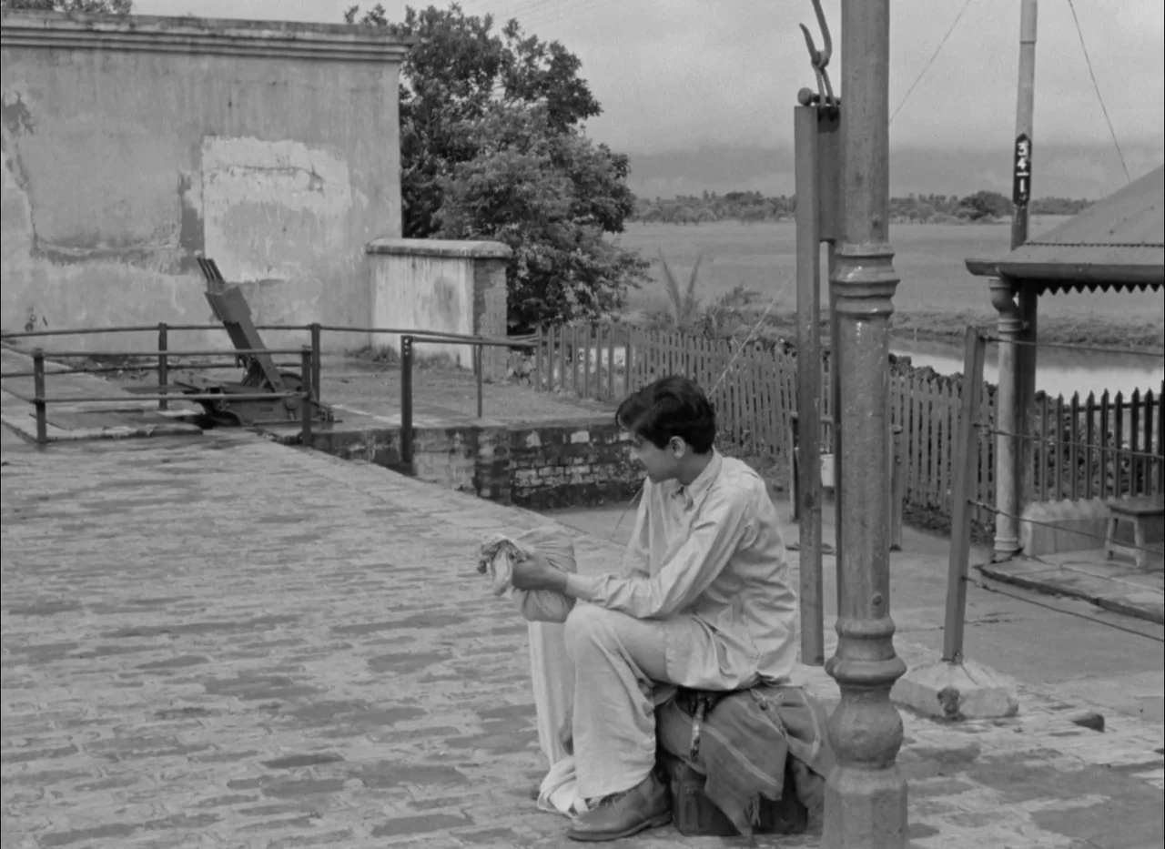 Aparajito (1956) movie still: fullbody — A rural train station platform features a stone floor and a metal pole. The young man sits on a small piece…