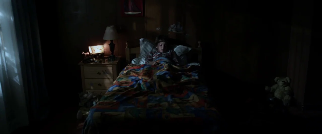 Insidious (2010) movie still: wide — A dark, wood-paneled room contains a child sleeping in a single bed under a quilt. A small table lamp sits on a…