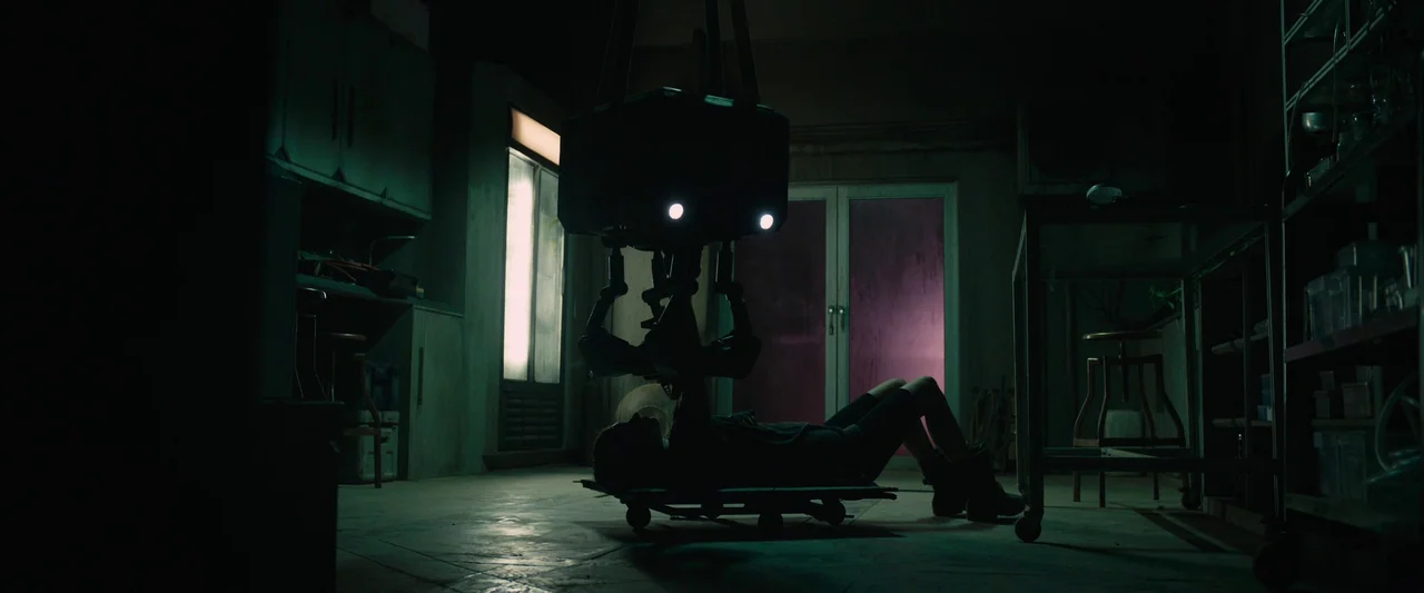 Settlers (2021) movie still: wide — A dimly lit industrial workspace contains cluttered shelves and dark, metallic surfaces. A lone technician lies…