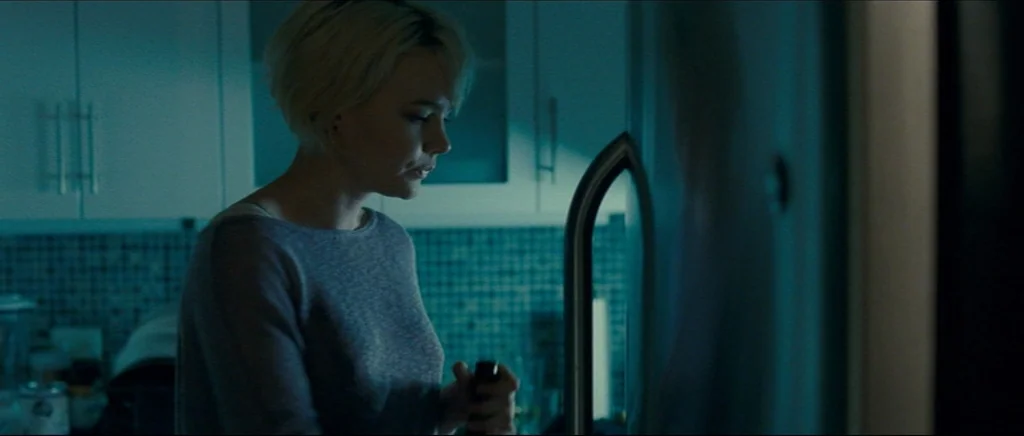 Shame (2011) movie still: medium — A dimly lit room with blue-tinted shadows and mosaic tiled backsplash. A woman in a grey sweater stands before an…