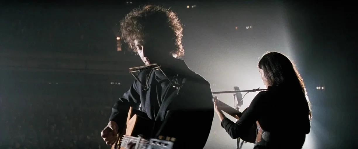 A Complete Unknown (2024) movie still: medium — A dark concert stage is illuminated from behind by a harsh spotlight. A man playing a guitar and…