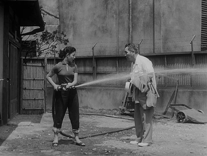I Live In Fear (1955) movie still: fullbody — A gravel yard with a bamboo fence and a concrete wall in the background. A young woman directs a water…