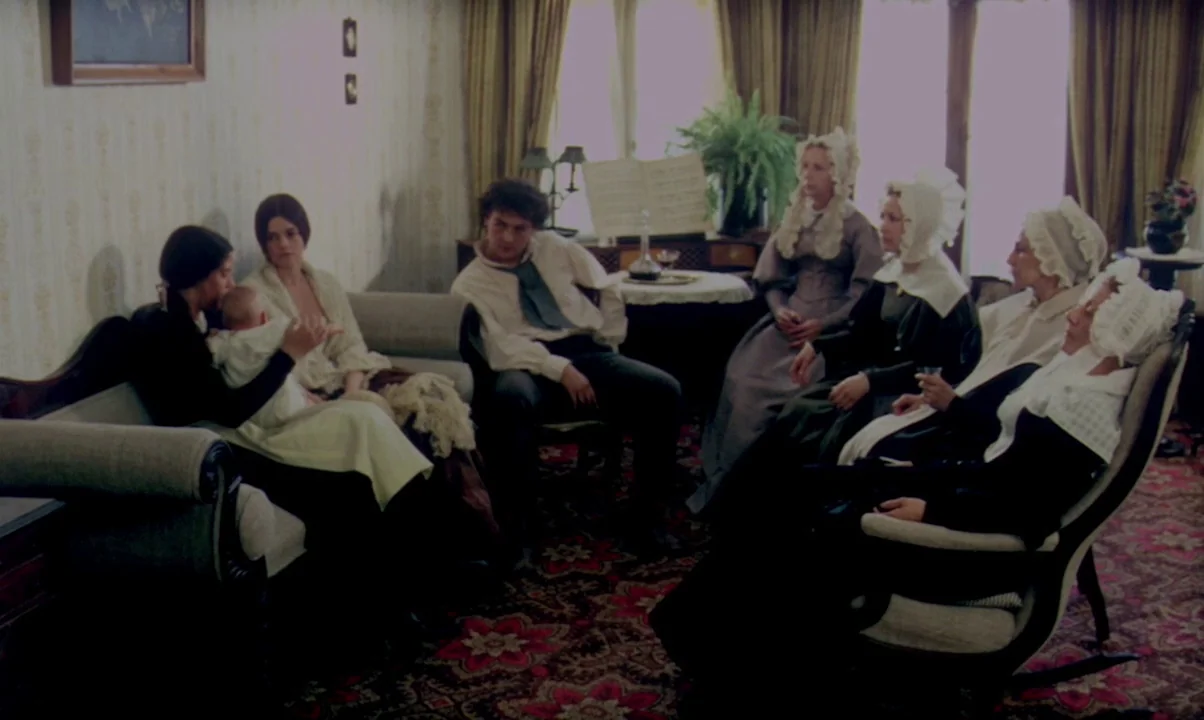 Kamouraska (1973) movie still: wide — A 1900s living room features patterned wallpaper and a heavily detailed rug. Several people sit together on…
