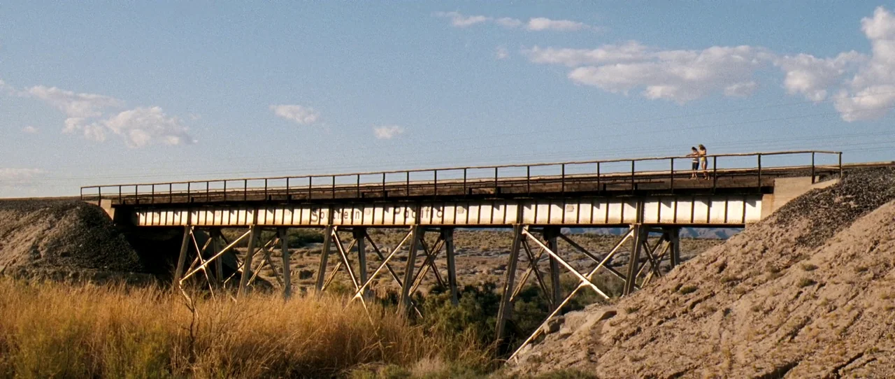 Little Birds (2011) movie still: wide — A long metal Southern Pacific railway bridge spans a dry, arid desert landscape. Two young women stand on…