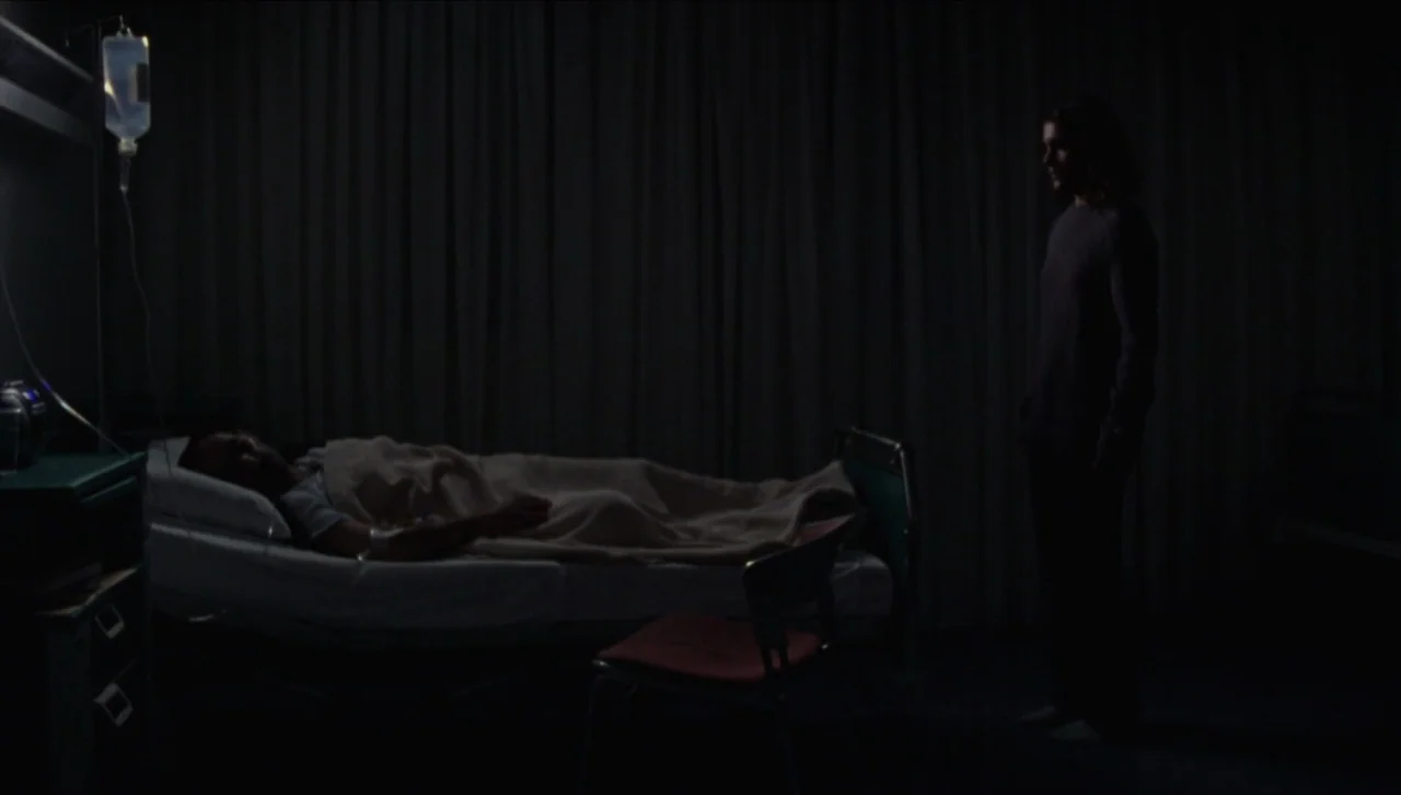 Attenberg (2010) movie still: wide — A dark hospital room contains a patient lying in a bed with IV fluids. A second man stands silently near the…