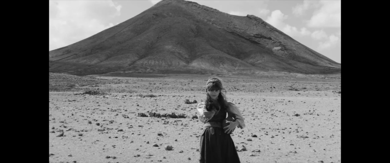 The Man Who Killed Don Quixote (2018) movie still: fullbody — A woman stands alone in a vast, arid volcanic desert beneath a large mountain. She…