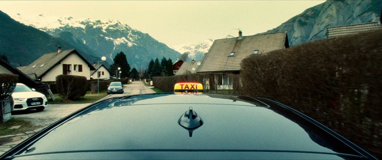 The Five Devils (2022) movie still: wide — A view from the roof of a moving taxi traveling down a suburban road. Houses with hedges and snow-capped…