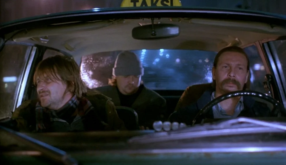 Night on Earth (1991) movie still: medium — The interior of a moving taxi at night with city lights blurred through windows. Three men sit in the…