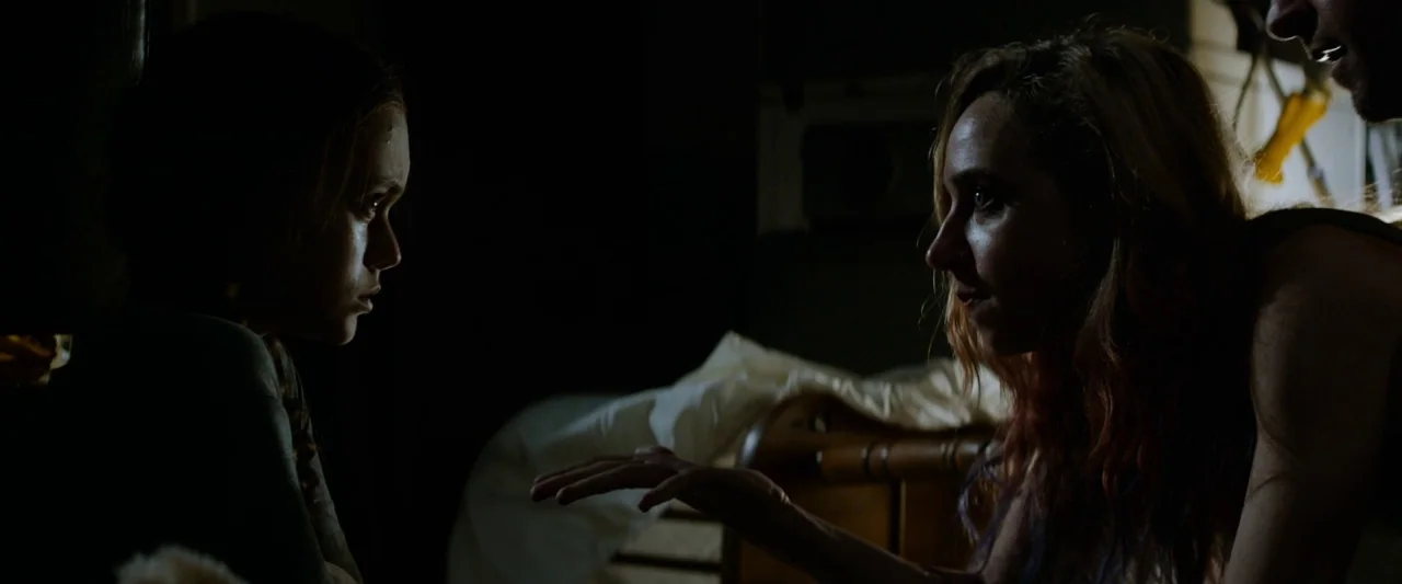 The Monster (2016) movie still: medium — A dark, dimly lit indoor space with a wooden bed frame visible behind the subjects. Two young women face…