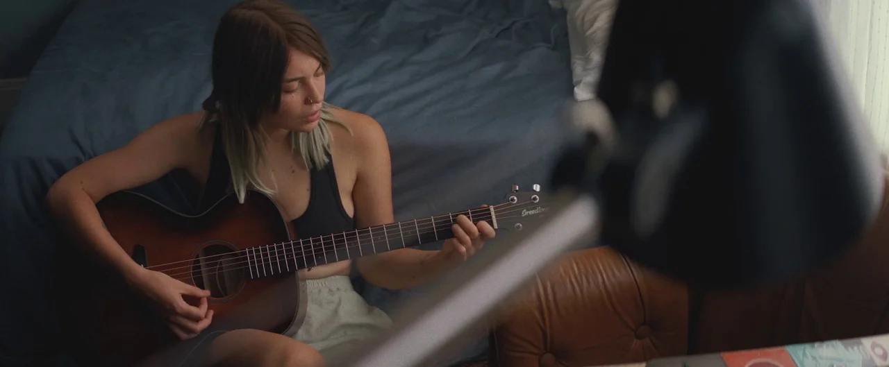 Poser (2021) movie still: medium — A person sits on the floor of a bedroom against a rumpled blue bed cover. The individual is playing a brown…
