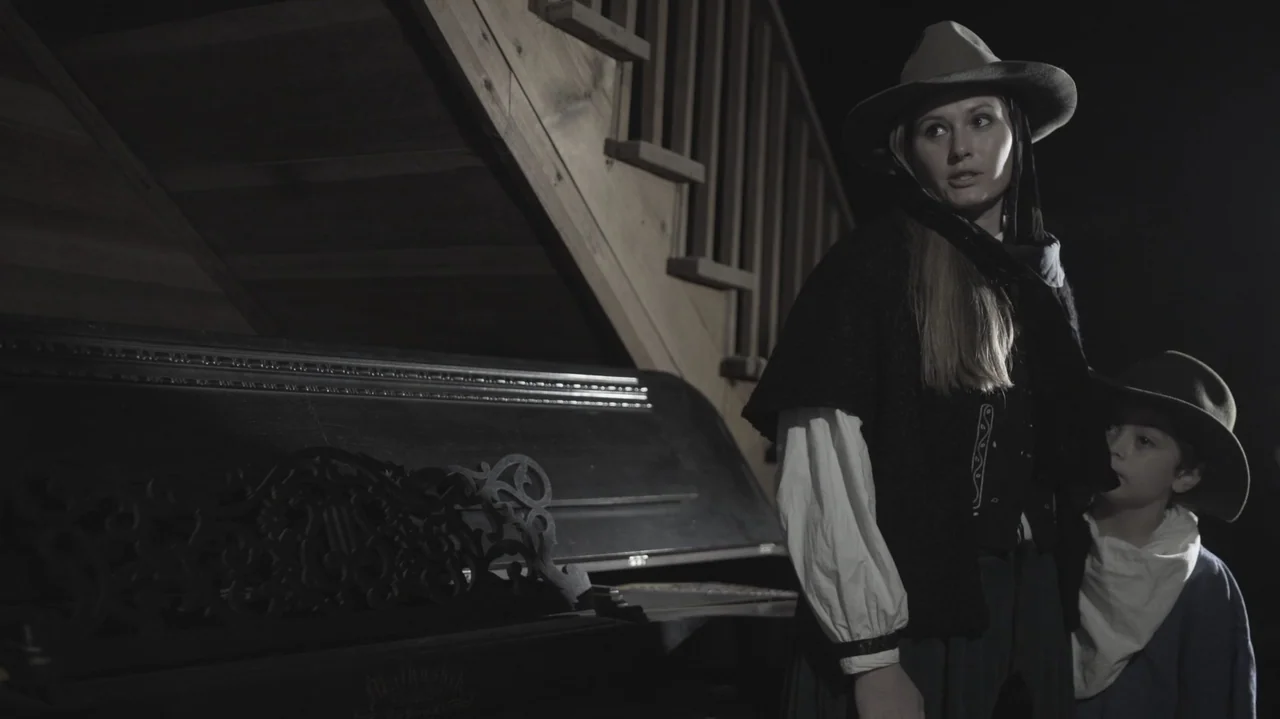 The Dalton Gang (2020) movie still: medium — A dark room features a wooden staircase in the back and an upright piano. A woman and a young boy stand…