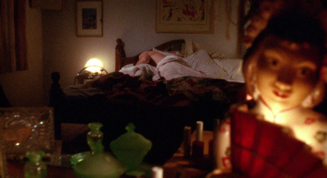White Of The Eye (1987) movie still: medium — A dimly lit room with a person sleeping in a bed in the background. A figurine stands in the…