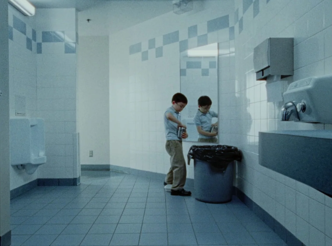Riceboy Sleeps (2022) movie still: wide — A public tiled restroom with white walls and blue accent squares. A young boy stands by the mirror near a…