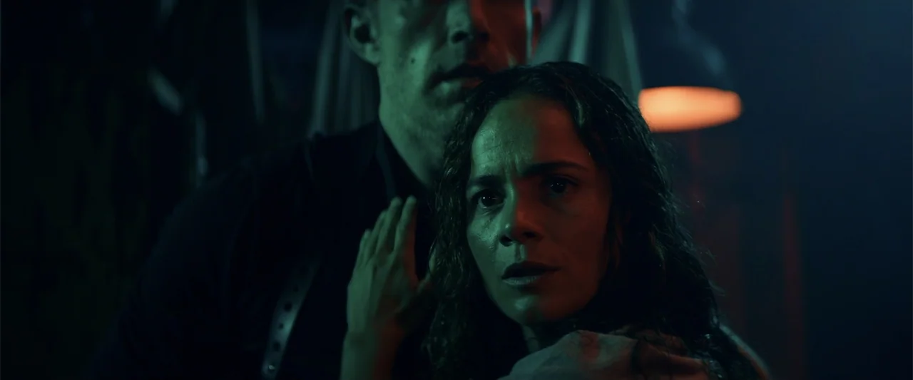 Hypnotic (2023) movie still: medium — A dark, dimly lit industrial space features a man standing behind a woman. The woman stares forward with a…