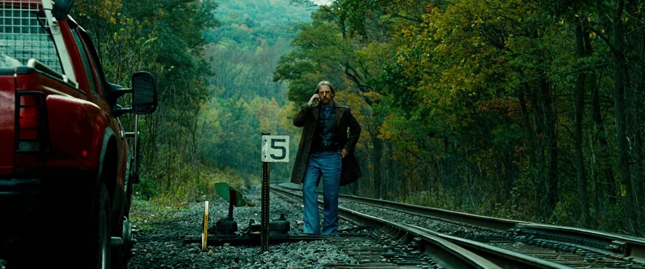 Unstoppable (2010) movie still: wide — A man stands between sets of tracks in a dense autumn forest setting. He holds a phone to his ear while…